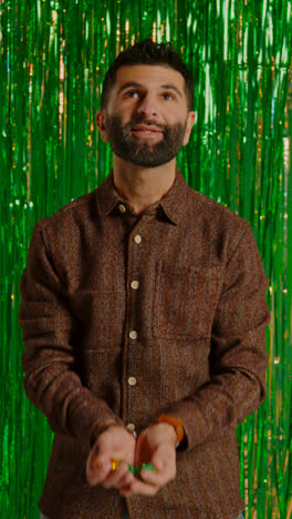 Vertical-Video-Of-Man-Celebrating-St-Patrick's-Day-Standing-In-Front-Of-Green-Tinsel-Curtain-And-Throwing-Novelty-Gold-Coins-In-The-Air